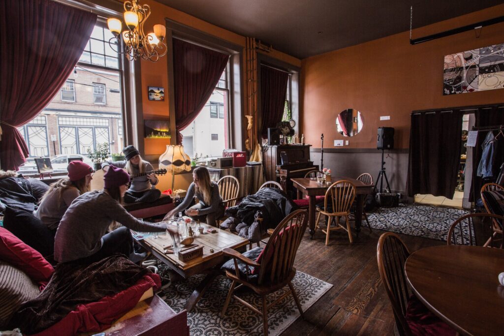 A cozy room of a restaurant. There are wooden tables with chairs across the room. Four individuals are sitting together; one of them is playing guitar. Near the entrance of the room, there is a stage with a piano and a speaker.