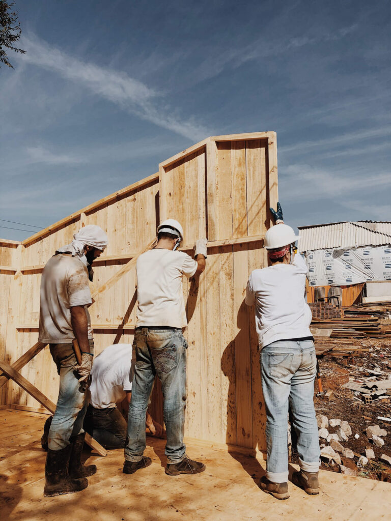 Three men in white t-shirts and jeans and white hardhats constructing a fence outside. 