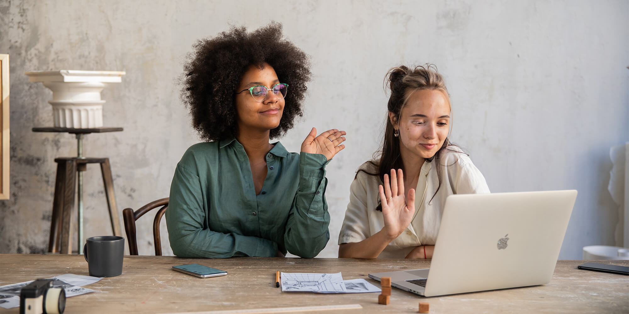 two women sitting at a desk wave at an open laptop 