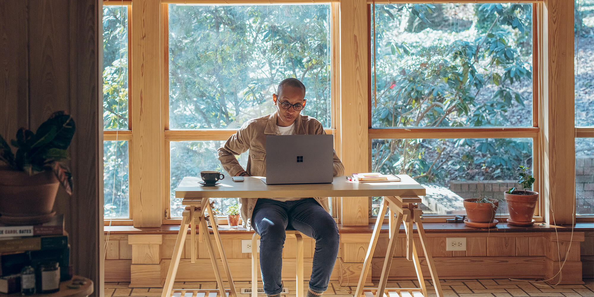 A man sits at a desk in front of a laptop, with open windows behind him revealing a forest.